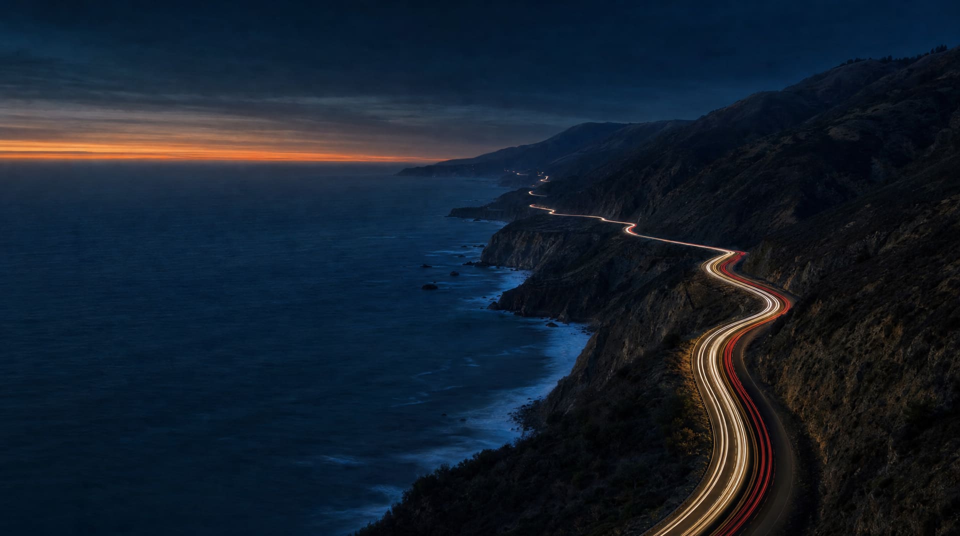 Aerial view of a winding coastal highway at dusk, traced by light trails, with the ocean to the left.